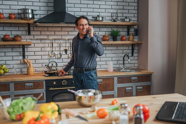 Vrolijke man staan op fornuis en praten over de telefoon in de keuken. Hij kijkt terug en glimlacht. Laptop en coloful groenten en kruiden op tafel.