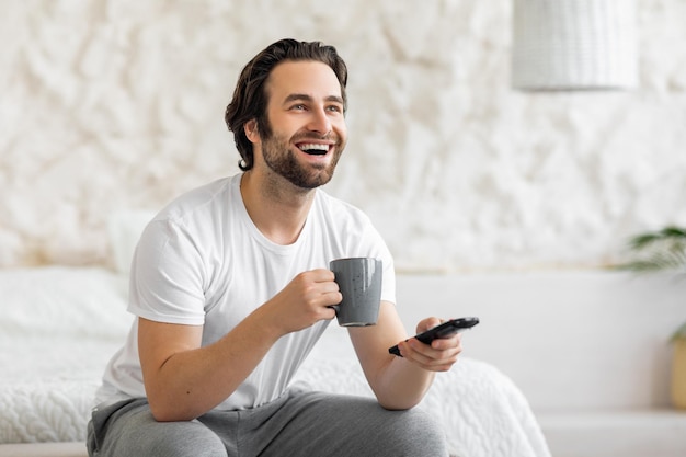 Vrolijke man zittend op bed tv kijken en koffie drinken