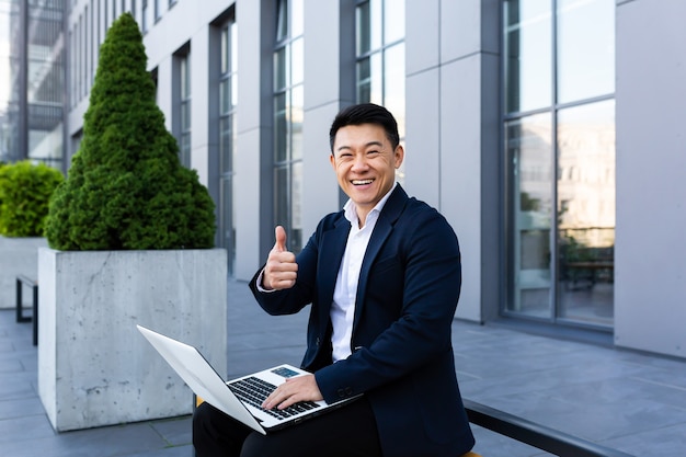 Vrolijke mannelijke aziatische zakenman die aan laptop online bankieren op computer in de buurt van kantoorcentrum werkt en naar de camera kijkt en glimlacht
