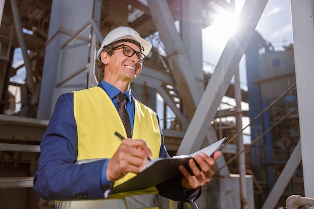 Vrolijke mannelijke ingenieur die documenten buiten schrijft in de fabriek