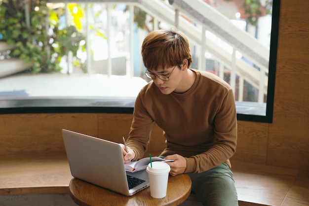 Vrolijke mannelijke journalist in trendy bril, graag klaar met het afronden van het werk aan het herlezen van boeken, geschreven in notitieboekjetekst voordat hij naar de redacteur belt om de publicatie te presenteren die koffie drinkt voor de lunch in café