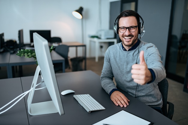 Vrolijke mannelijke technologieondersteuningsmanager die duimen in bureau tonen.