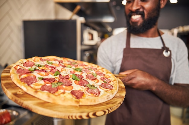 Foto vrolijke mannelijke werknemer in schort kijken naar houten plank met pizza en glimlachen