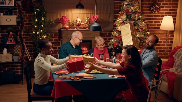 Vrolijke mensen verzamelden zich aan de kersttafel terwijl ze geschenken uitwisselden. Multi-etnische familieleden vieren traditionele wintervakantie terwijl ze elkaar cadeautjes geven.