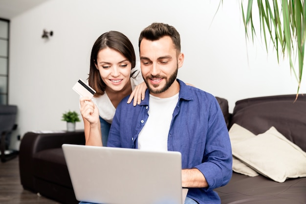 Vrolijke moderne jonge paar in casual kleding zittend op de Bank en met behulp van laptop en onbeperkte creditcard tijdens het samen online winkelen