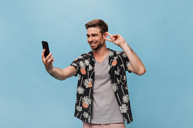 Vrolijke moderne man met cool kapsel in zomershirt met korte mouwen die selfie maakt met vredesteken en glimlachend op blauwe achtergrond