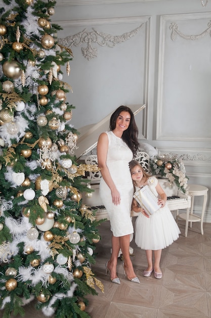 Vrolijke moeder en haar schattige dochter meisje met een witte piano en versierde kerstboom met cadeautjes met plezier en knuffelen in klassieke luxe Nieuwjaar interieur.