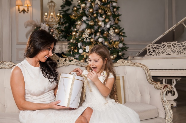 Vrolijke moeder en haar schattige dochter meisje uitwisselen van geschenken in wit klassiek interieur tegen de achtergrond van een piano en een versierde kerstboom.