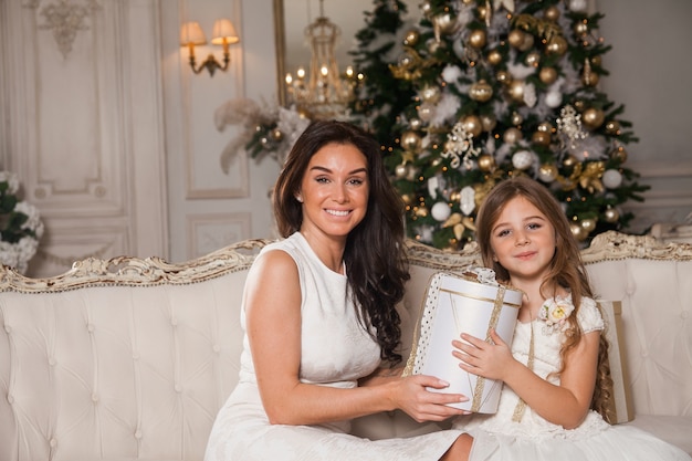 Vrolijke moeder en haar schattige dochter meisje uitwisselen van geschenken in wit klassiek interieur tegen de achtergrond van een piano en een versierde kerstboom.