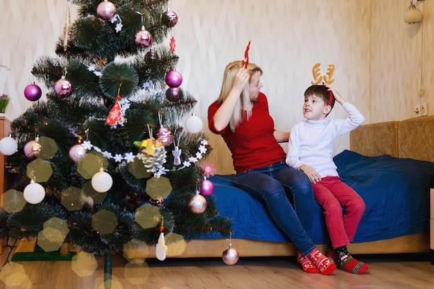 Vrolijke moeder en haar zoon, kleine jongen die plezier heeft. Kerstboom bij gezellig huis. truien, hertenhoorns