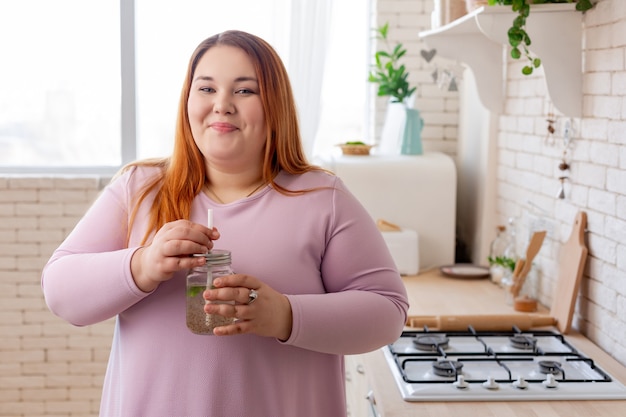 Vrolijke mollige vrouw die naar je lacht terwijl ze een gezonde smoothie drinkt