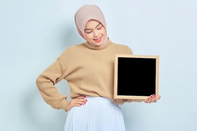 Vrolijke mooie aziatische moslimvrouw in bruine trui met schoolbord geïsoleerd op witte achtergrond mensen religieuze levensstijl concept