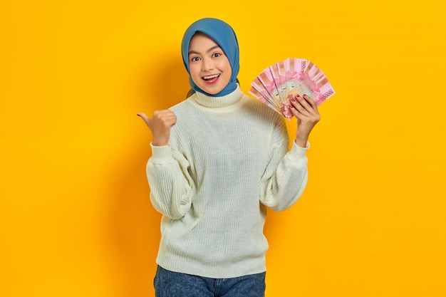 Vrolijke mooie Aziatische moslimvrouw in witte trui duimen wijzend op lege ruimte met contant geld in Indonesische rupiah bankbiljetten over gele achtergrond Mensen religieuze levensstijl concept