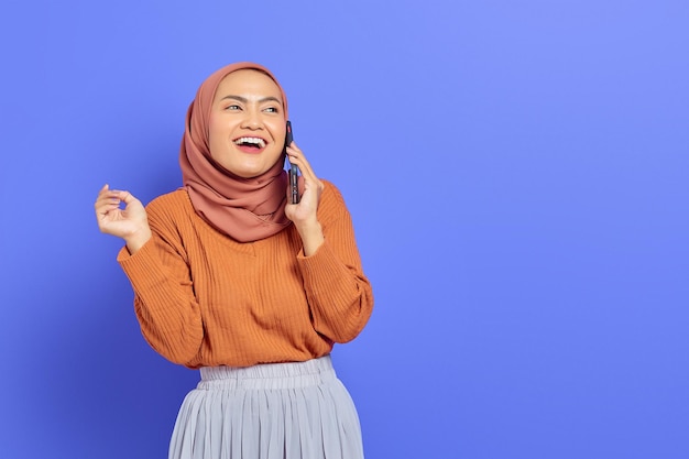 Vrolijke mooie Aziatische vrouw in bruine trui en hijab praten op mobiele telefoon terwijl ze opzij kijkt geïsoleerd over paarse achtergrond. Mensen islam religieus concept