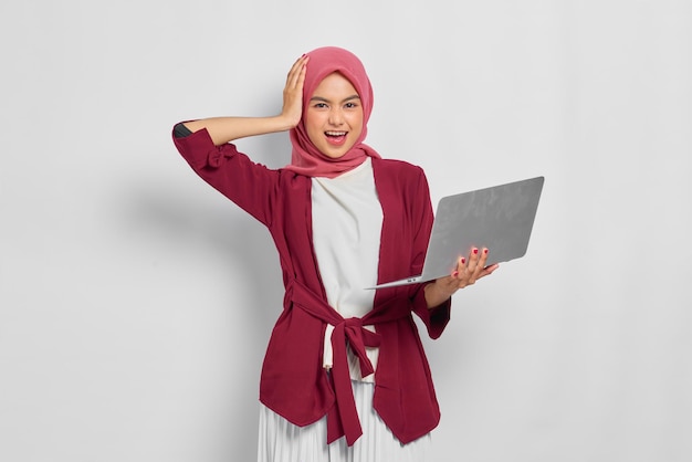 Vrolijke mooie Aziatische vrouw in casual shirt en hijab met een laptopcomputer, hand op het hoofd geïsoleerd op witte achtergrond. Mensen religieuze levensstijl concept