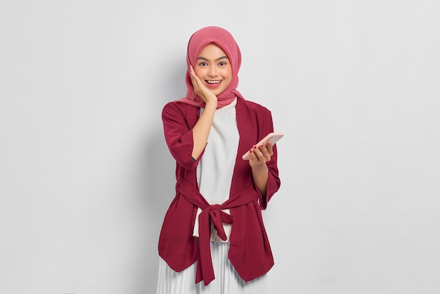 Vrolijke mooie Aziatische vrouw in casual shirt en hijab met mobiele telefoon geïsoleerd op witte achtergrond. Mensen religieuze levensstijl concept