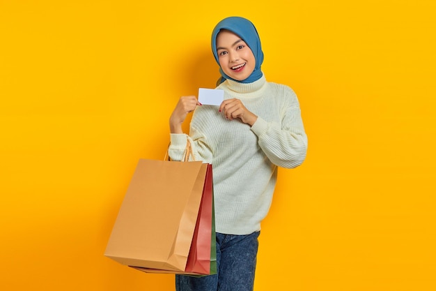 Vrolijke, mooie Aziatische vrouw in een witte trui die boodschappentassen vasthoudt en een creditcard toont die over een gele achtergrond is geïsoleerd