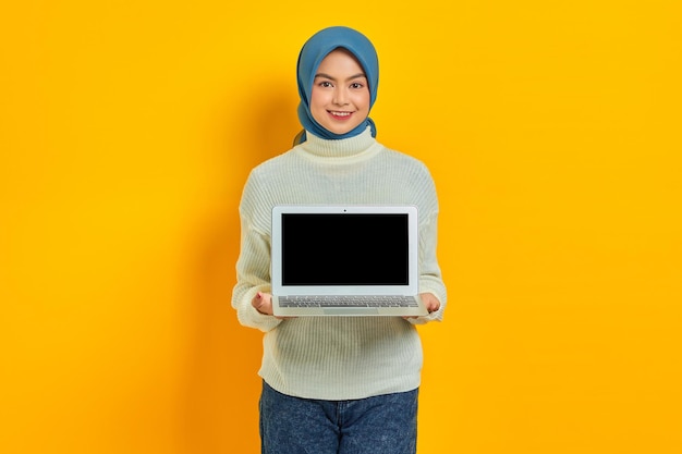Vrolijke mooie Aziatische vrouw in witte trui en hijab met een laptop met leeg scherm geïsoleerd over gele achtergrond mensen religieuze levensstijl concept