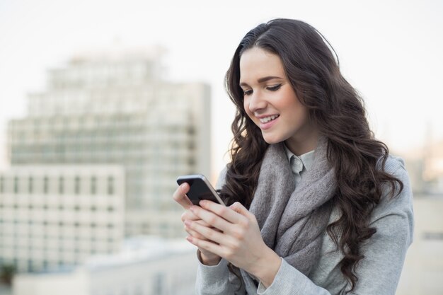 Vrolijke mooie brunette in winterkleren die een tekst op haar smartphone verzenden