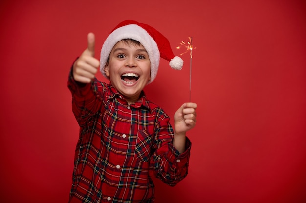 Vrolijke mooie geweldige preteen jongen in kerstmuts en geruit rood shirt houdt Bengaalse kerstverlichting, sterretjes in zijn handen, toont duim omhoog staat tegen een gekleurde achtergrond met kopieerruimte voor advertentie