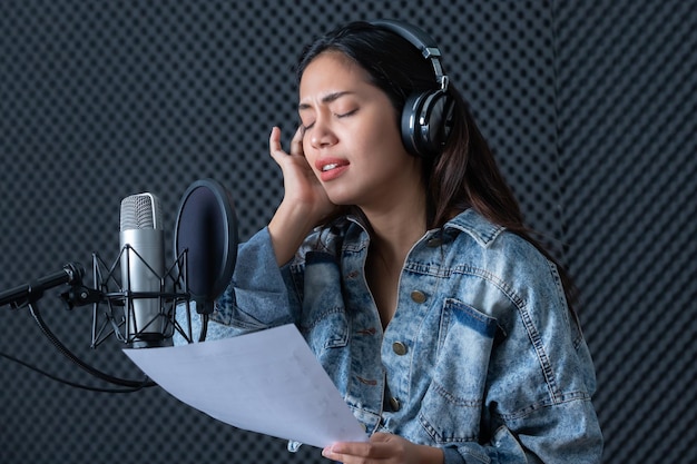 Vrolijke, mooie glimlach van het portret van een jonge Aziatische zangeres die een koptelefoon draagt die een nummer van de microfoon opneemt in een professionele studio