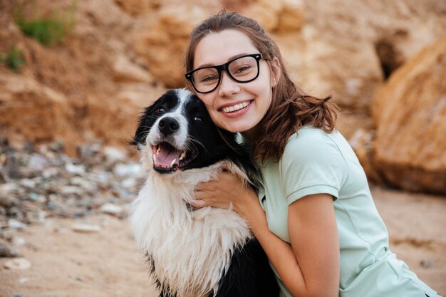 Vrolijke mooie jonge vrouw die haar hond op het strand zit te knuffelen