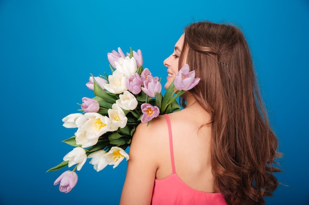 Vrolijke mooie jonge vrouw die lacht en een boeket lentebloemen vasthoudt over blauwe achtergrond