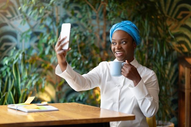 Vrolijke mooie jonge zwarte vrouw die in café zit en selfie neemt