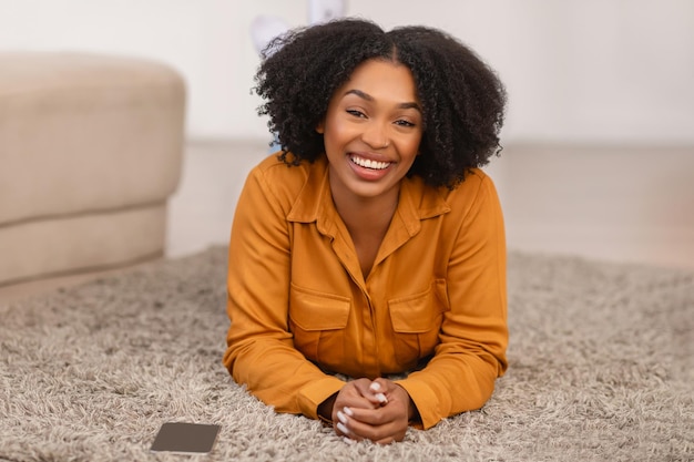 Vrolijke mooie millennial krullende Afrikaans-Amerikaanse vrouw ligt op de vloer met telefoon