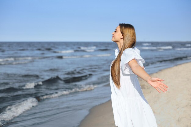 Vrolijke, mooie vrouw op het oceaanstrand in een witte zomerjurk, open armen.