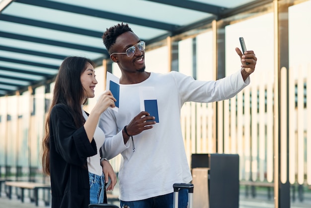 Vrolijke multiraciale mannelijke en vrouwelijke vrienden selfie maken op smartphone met paspoorten en tickets voor reis.