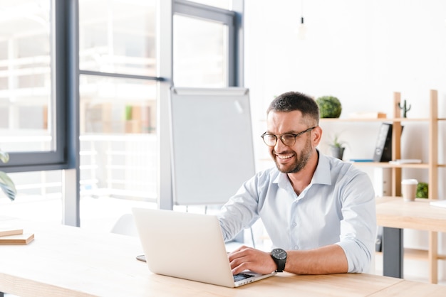 vrolijke office man 30s in wit overhemd zit aan bureau en werkt op laptop in business center