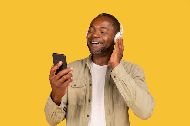 Vrolijke ontspannen volwassen Afro-Amerikaanse man in casual en draadloze koptelefoon kijkend naar telefoon