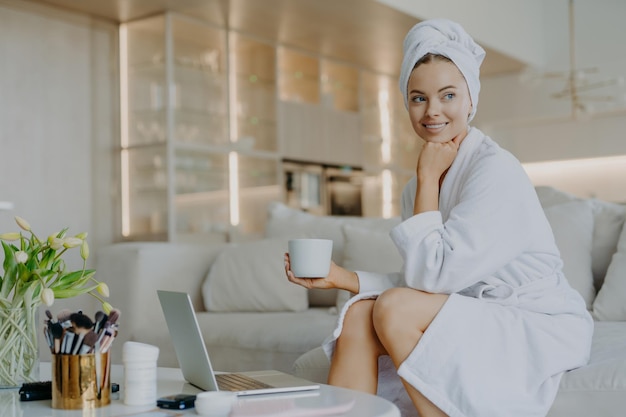 Vrolijke opgefriste vrouw met een gezonde huid draagt een badjas na het douchen drinkt koffie en kijkt bedachtzaam opzij zit op comfortabele bank werkt op laptopcomputer gebruikt cosmetische producten