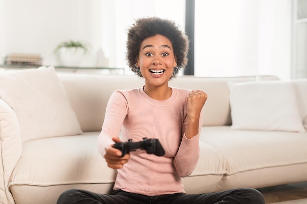 Vrolijke opgewonden jonge dame van gemengd ras met joystick die computerspel speelt, maakt overwinning en succes gebaar