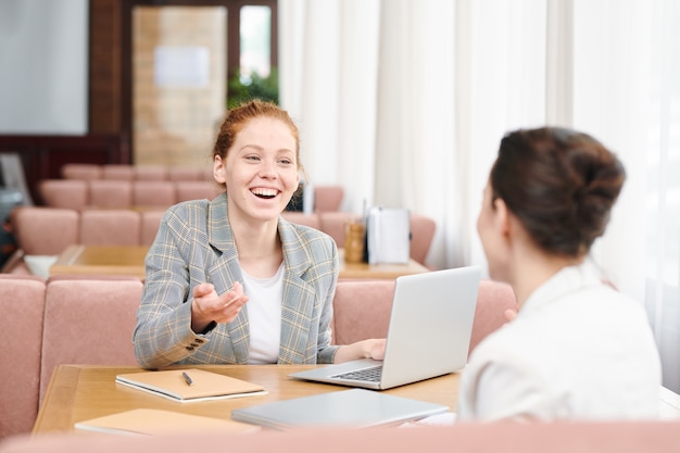 Vrolijke opgewonden jonge dames zitten aan tafel in café en lachen terwijl ze vreemde ideeën van project bespreken