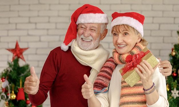 Vrolijke oude man en volwassen vrouw met kerstmutsen die met een glimlach naar de camera kijken en kerstcadeaus demonstreren tegen de muur