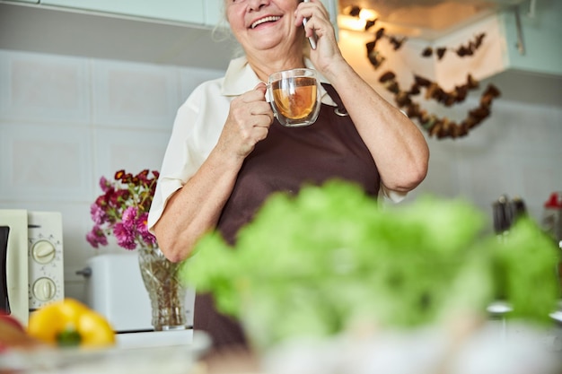 Vrolijke oude vrouw die op mobiel praat en thuis thee drinkt