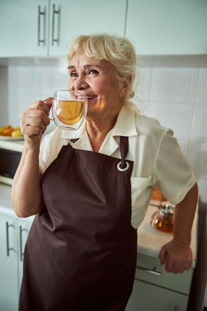 Vrolijke oude vrouw in schort die thuis thee drinkt