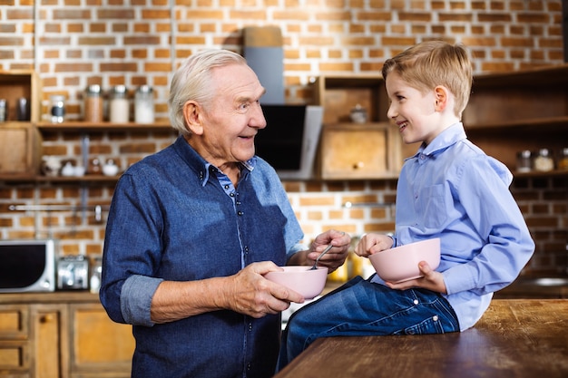 Vrolijke oudere man granen eten tijdens het ontbijt met zijn kleinzoon