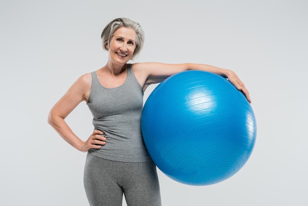 vrolijke oudere vrouw in sportkleding met blauwe fitnessbal geïsoleerd op grijs