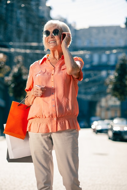 Vrolijke oudere vrouw met boodschappentassen bij zonnig weer glimlachend tijdens het telefoneren