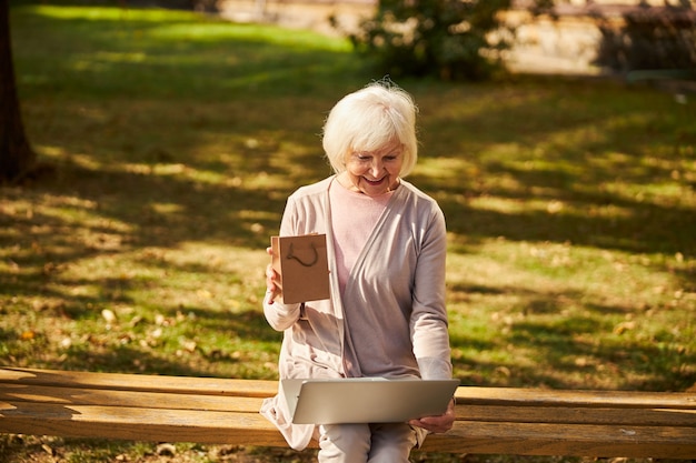 Vrolijke oudere vrouw op een bankje die op een zonnige dag met een laptop werkt en een klein pakketje vasthoudt