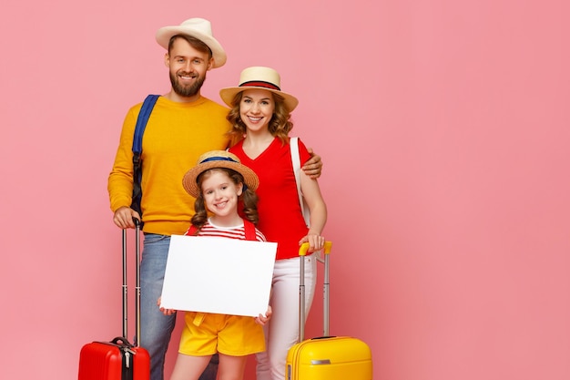 Vrolijke ouders en dochter met bagage en lege poster glimlachen en kijken naar camera tijdens zomervakantie tegen roze achtergrond