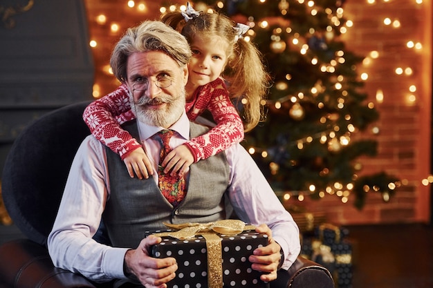 Vrolijke ouderwetse senior man met grijs haar en baard met een klein meisje met een geschenkdoos die elkaar omhelst.