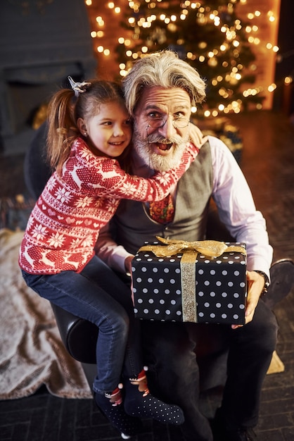 Vrolijke ouderwetse senior man met grijs haar en baard zitten met klein meisje in ingerichte kerstkamer.