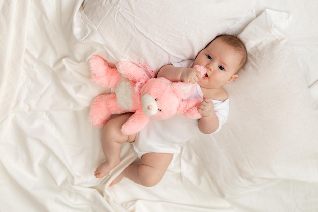 Vrolijke pasgeboren baby in een witte bodysuit ligt op een wit bed met een pluche roze speelgoedkonijn. producten voor kinderen, speelgoed. concept van een gelukkige jeugd en moederschap. kinderopvang. ruimte voor tekst.