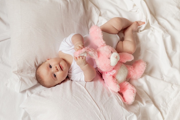 Vrolijke pasgeboren baby in een witte bodysuit ligt op een wit bed met een pluche roze speelgoedkonijn