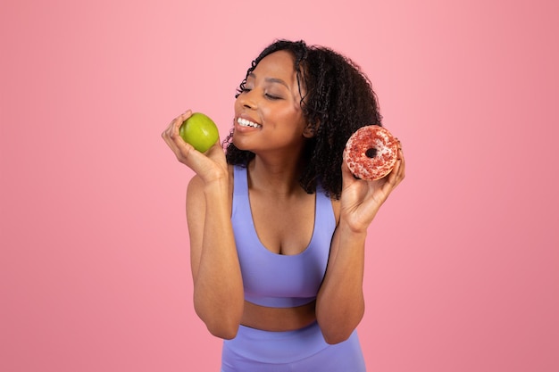 Vrolijke peinzende millennial afro-amerikaanse dame in sportkleding kiest donut of groene appel