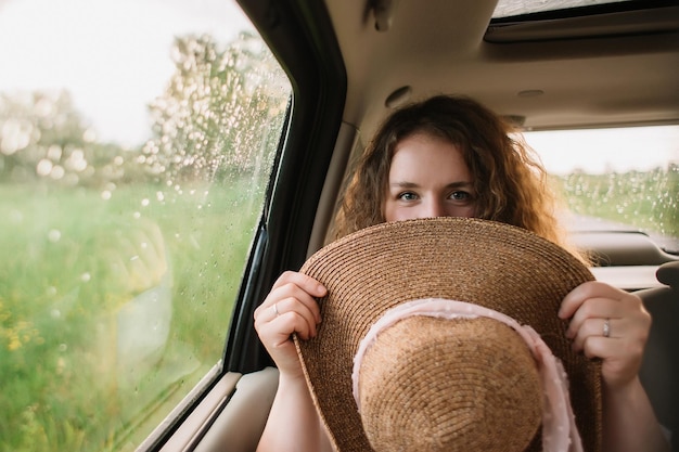 Vrolijke positieve gekrulde jonge vrouw in vrijetijdskleding zittend op de achterbank van de auto met hoed